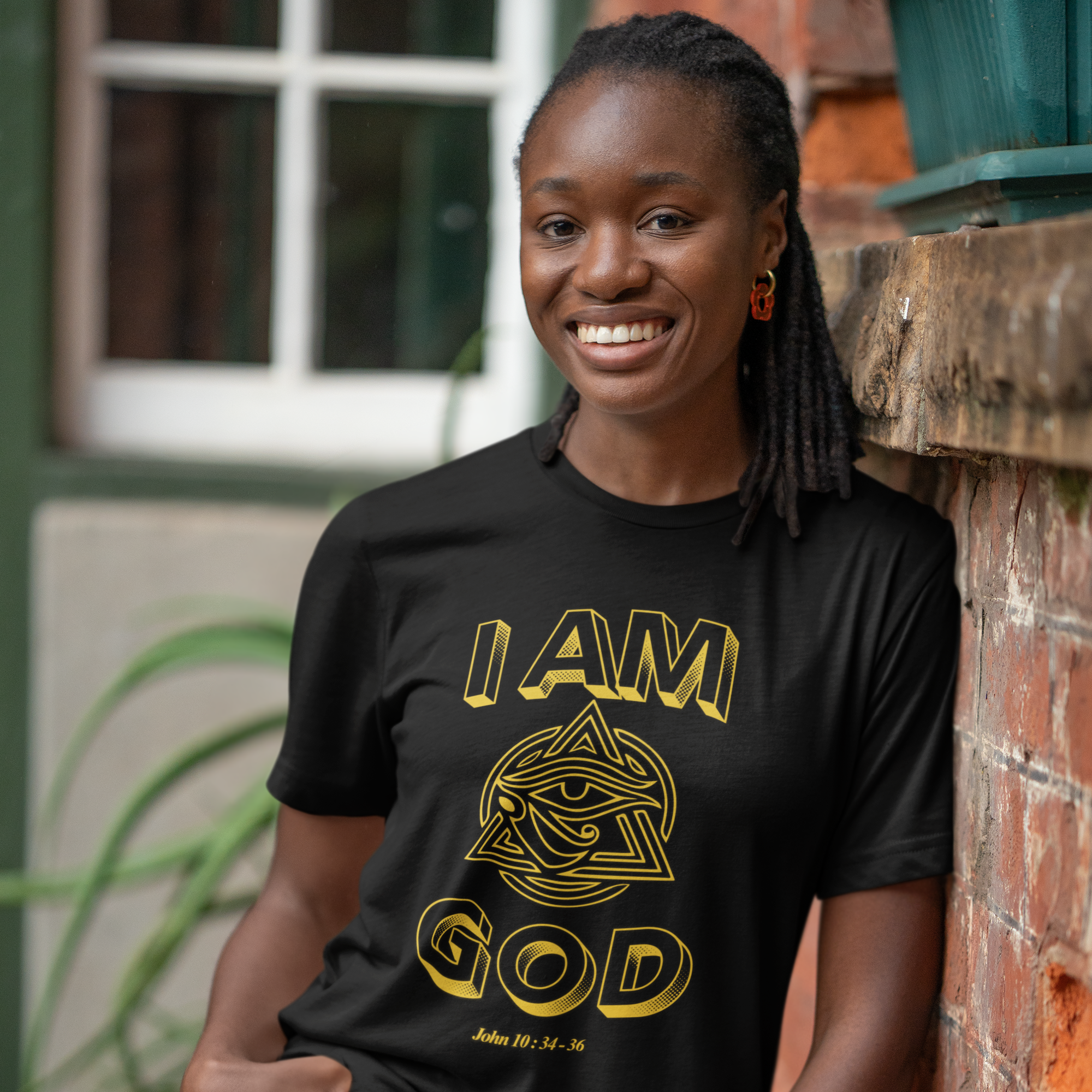 Woman wearing a black t-shirt with 'I AM GOD' printed in gold, standing against a brick wall.