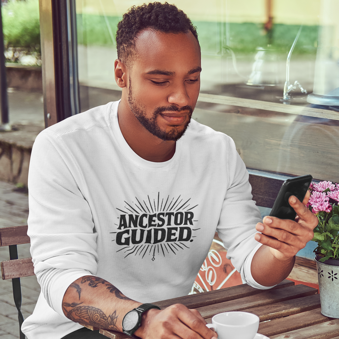 Man sitting at an outdoor cafe table using a phone with 'Ancestor Guided' shirt.