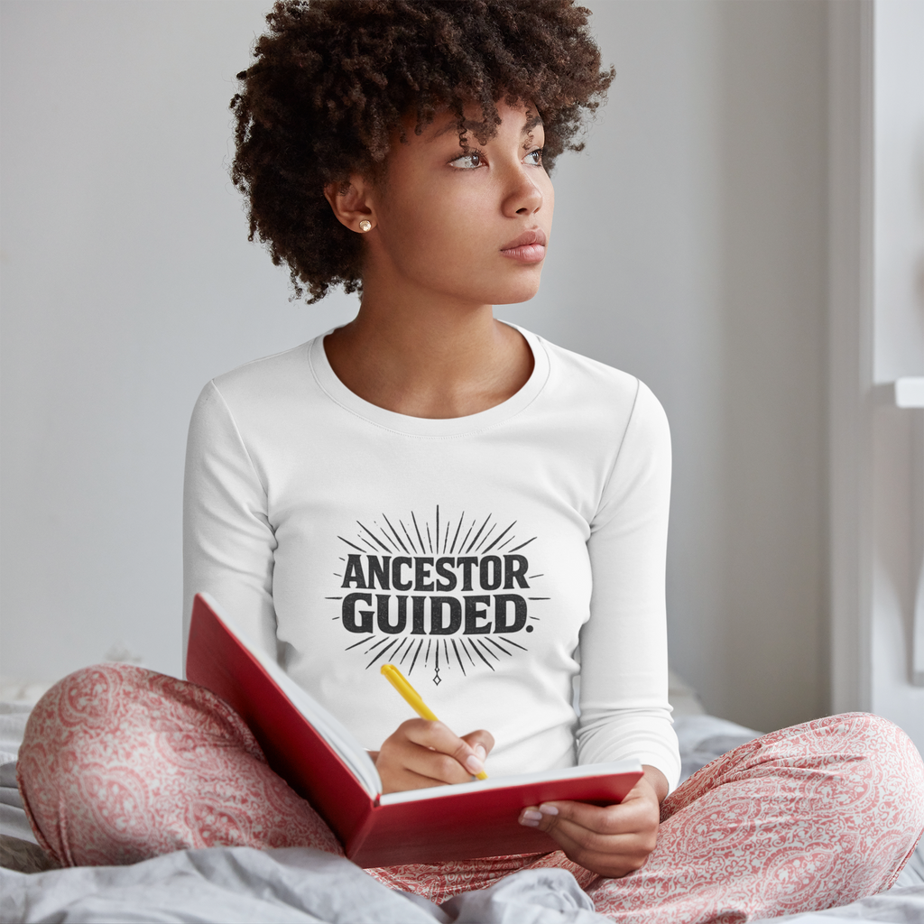 Person wearing a white long-sleeve shirt with 'ANCESTOR GUIDED' text, sitting on a bed holding a red notebook.